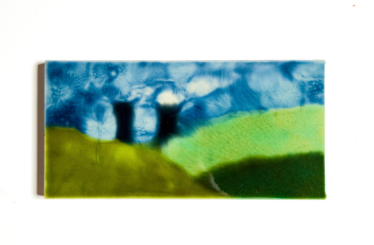 This image features a small glazed ceramic artwork titled "Cooling Tower." The artwork measures 3x6 inches and depicts an abstract landscape with rolling green hills under a vivid blue sky, accented by artistic representations of cooling towers in the distance.