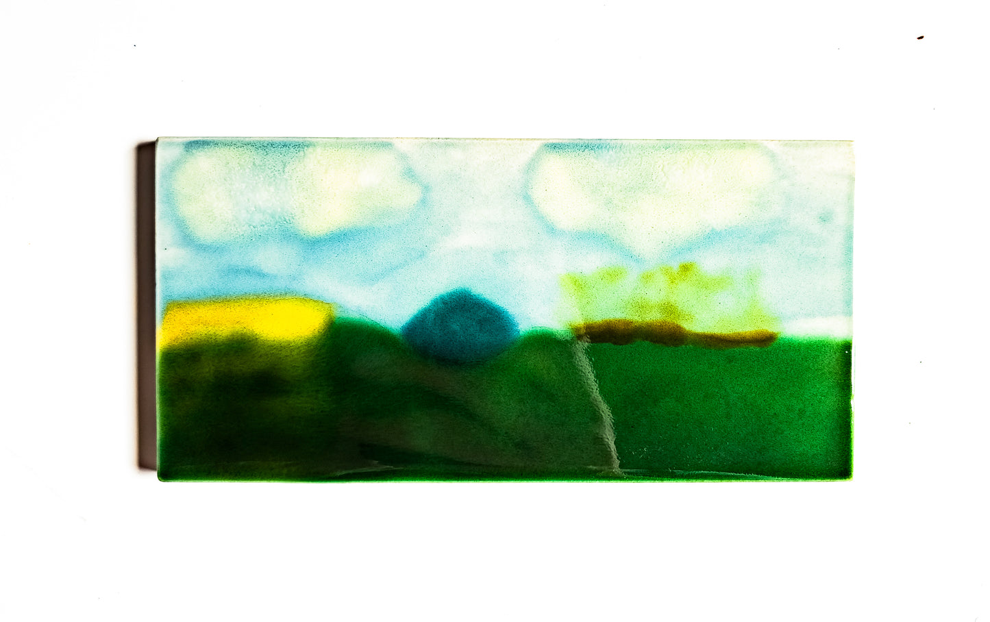 A glazed ceramic art piece depicting a simplistic landscape with green fields and blue sky.
